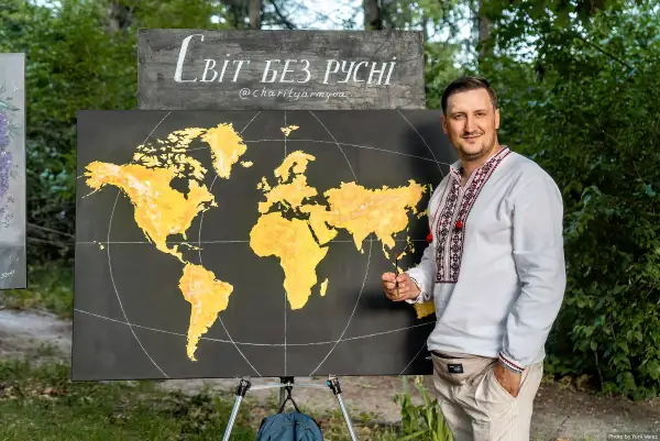 Чоловік у вишиванці стоїть біля картини з жовтою мапою світу та написом «Світ без русні».